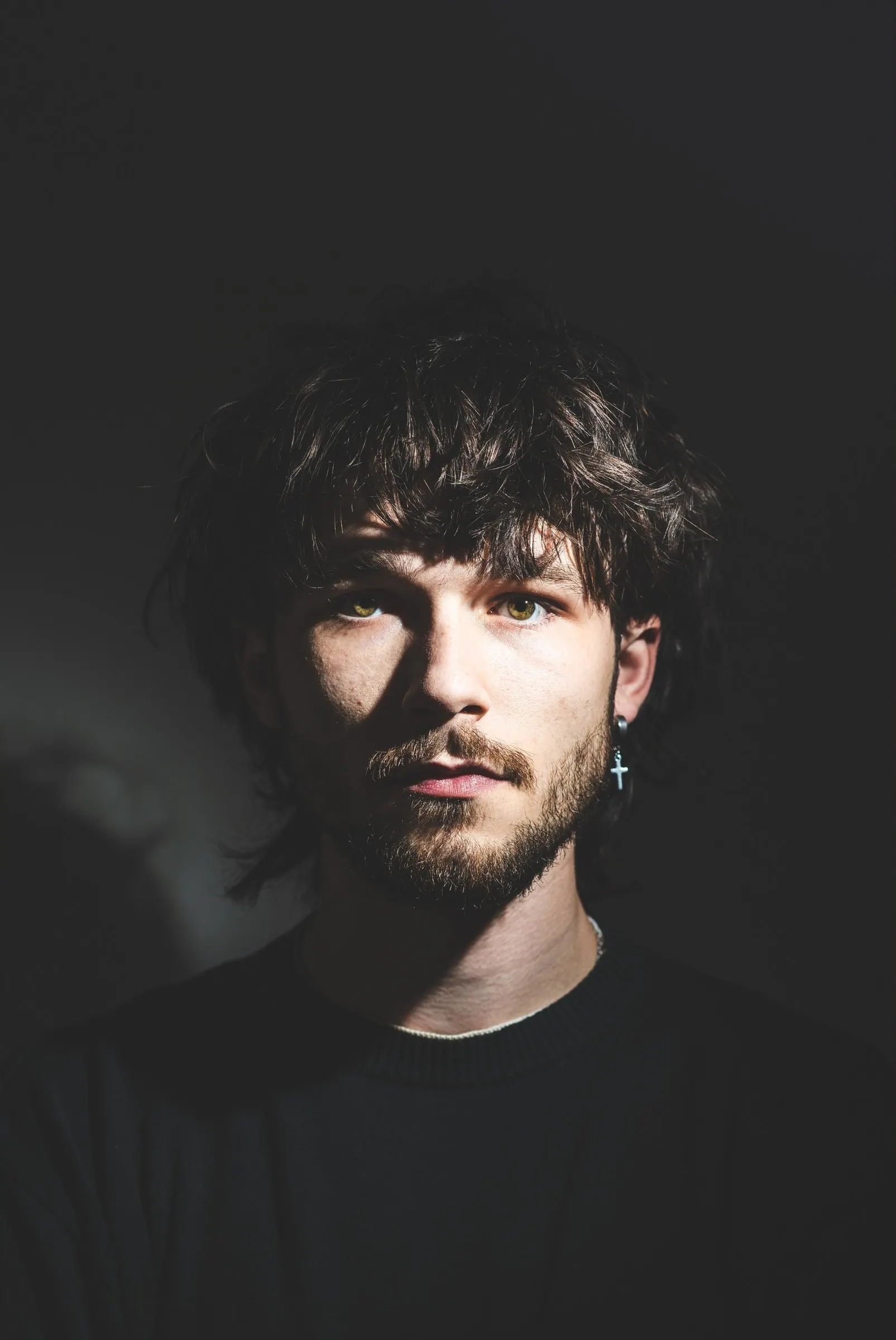 Portret van Joey, fotograaf uit Haaksbergen: donker warrig haar, baard en een kruisoorbel, met hard licht over zijn gezicht tegen een donkere achtergrond