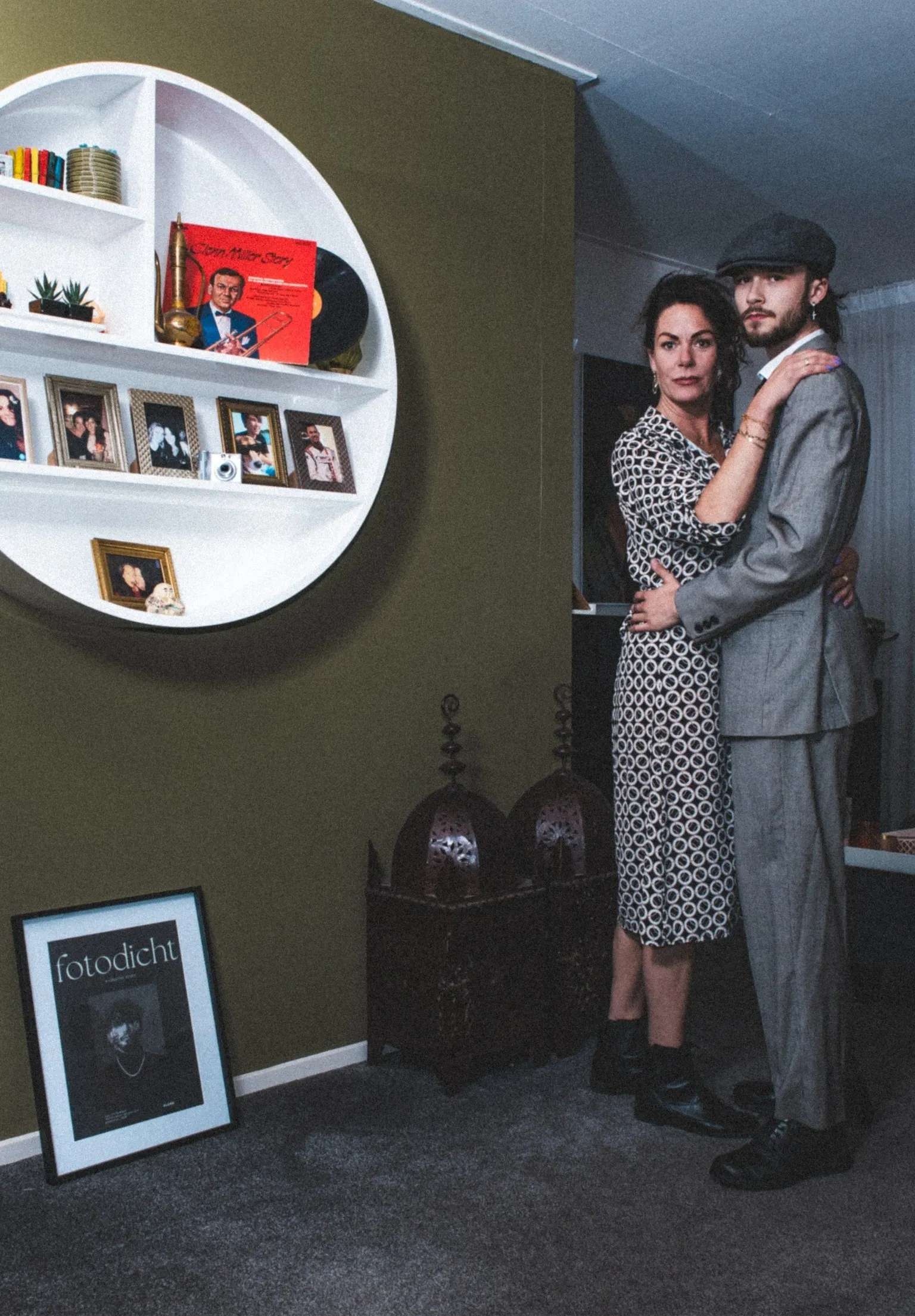 Stel in een woonkamer naast een rond wandrek met platen en fotolijstjes; zij in een zwart-witte jurk, hij in een grijs pak met pet