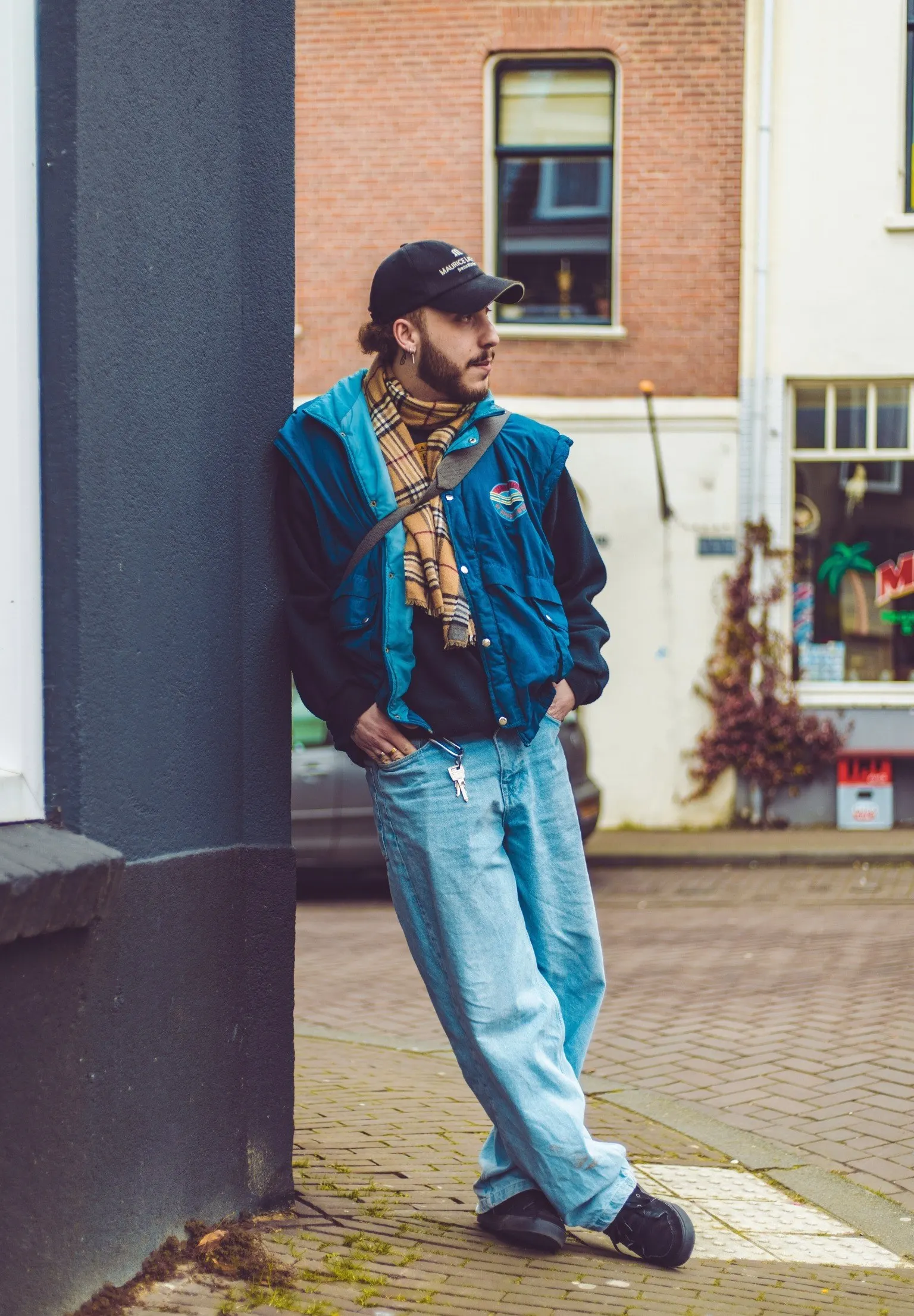 Jonge man met pet, geruite sjaal en blauwe retrojas leunt tegen een gevel in een straat met bakstenen huizen