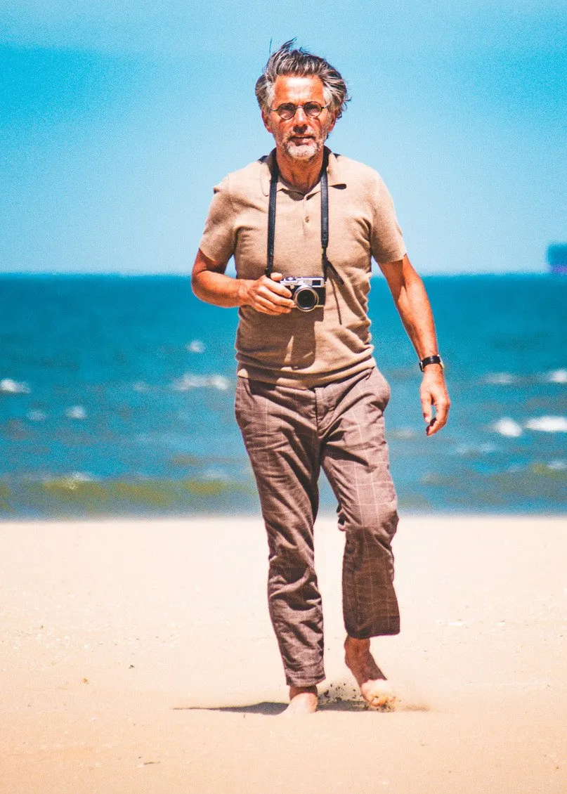 Man met grijs haar en een camera om zijn nek loopt op blote voeten over het strand, met de zee achter hem