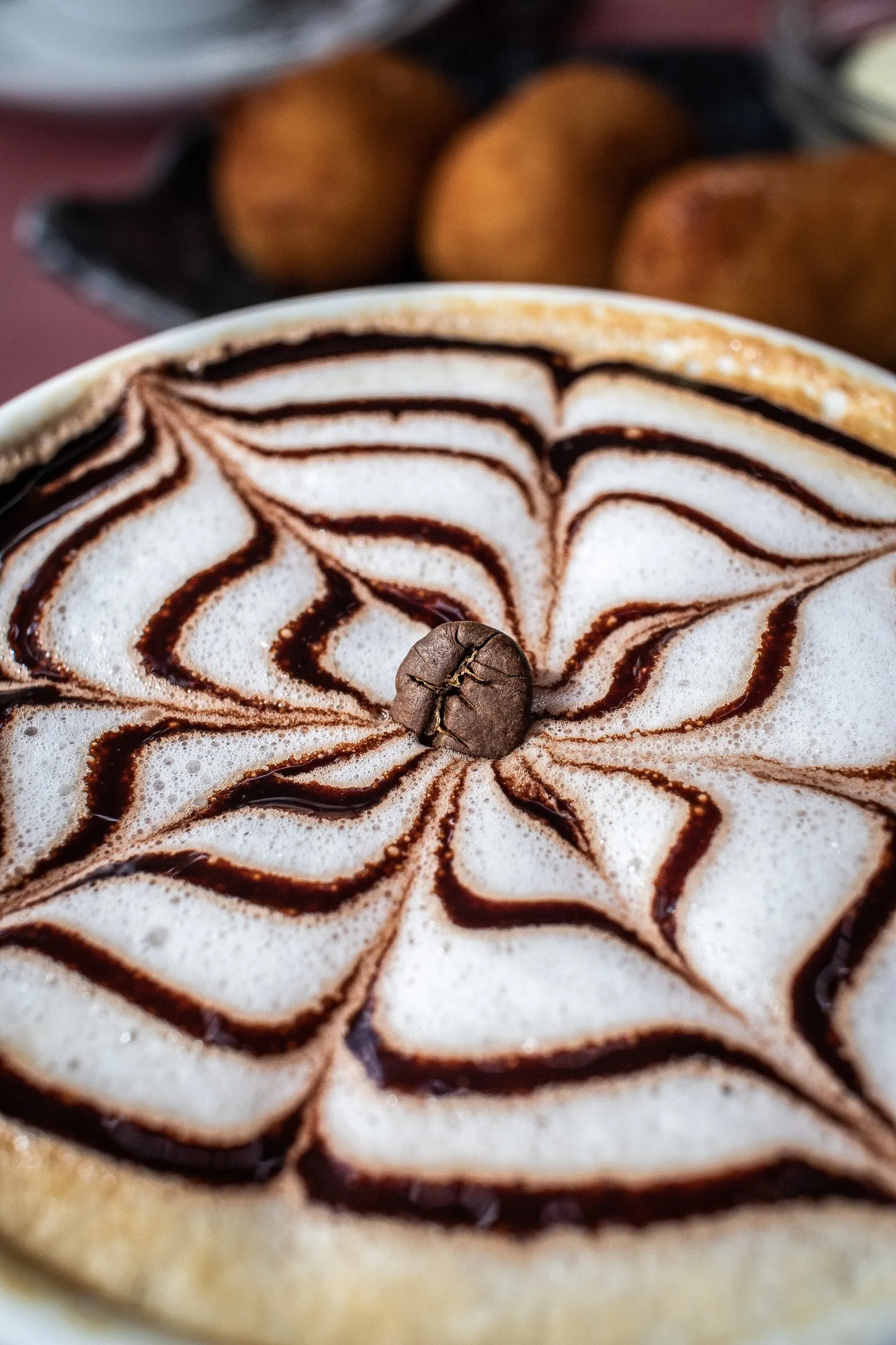 Cappuccino van bovenaf met een spinnenweb van chocoladesaus, met gebak op de achtergrond