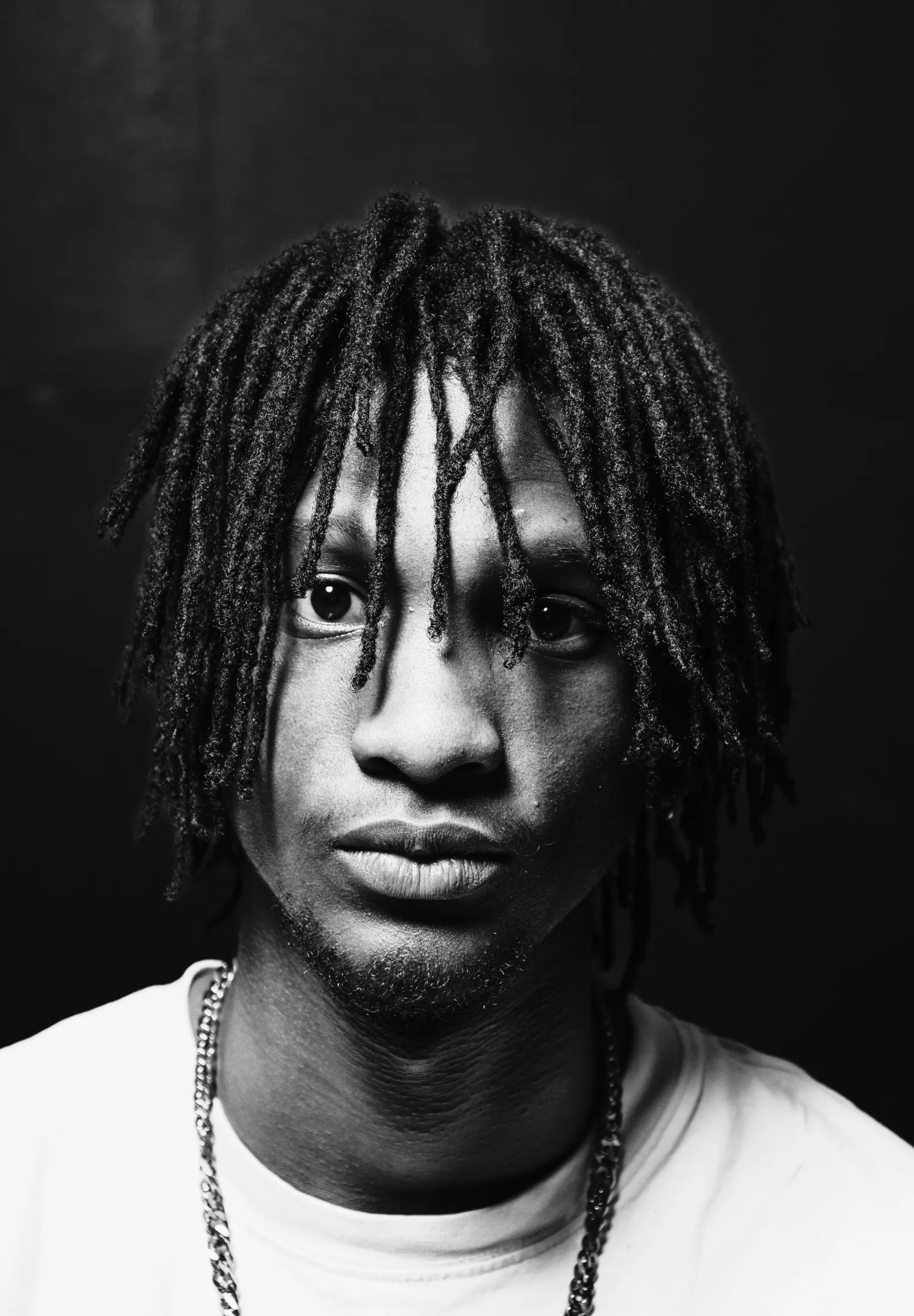 Zwart-witportret van een jonge man met dreadlocks en een wit shirt die recht in de camera kijkt