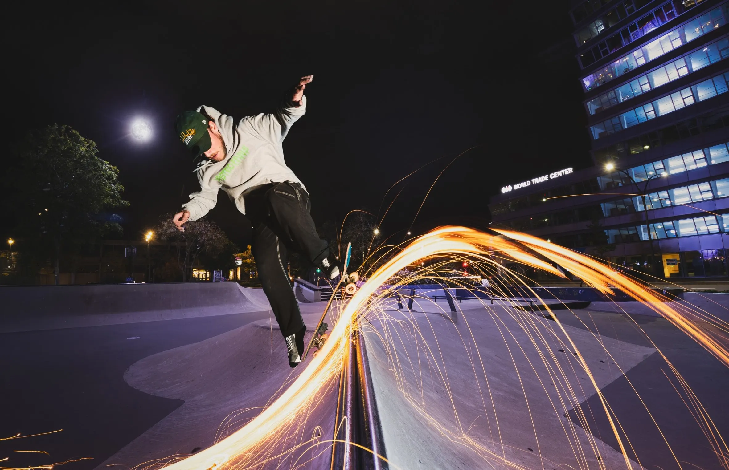 Skater springt 's nachts van een richel terwijl een spoor van vonken achter hem oplicht, met kantoorgebouwen op de achtergrond