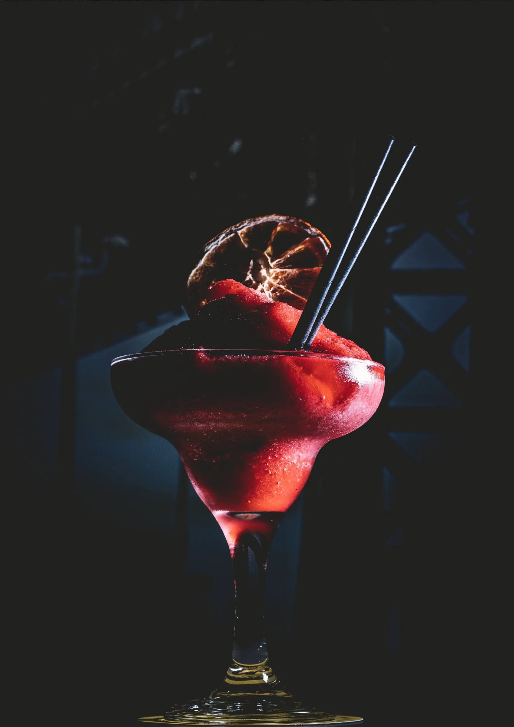Rode bevroren cocktail met een gedroogd sinaasappelschijfje en zwarte rietjes tegen een donkere achtergrond