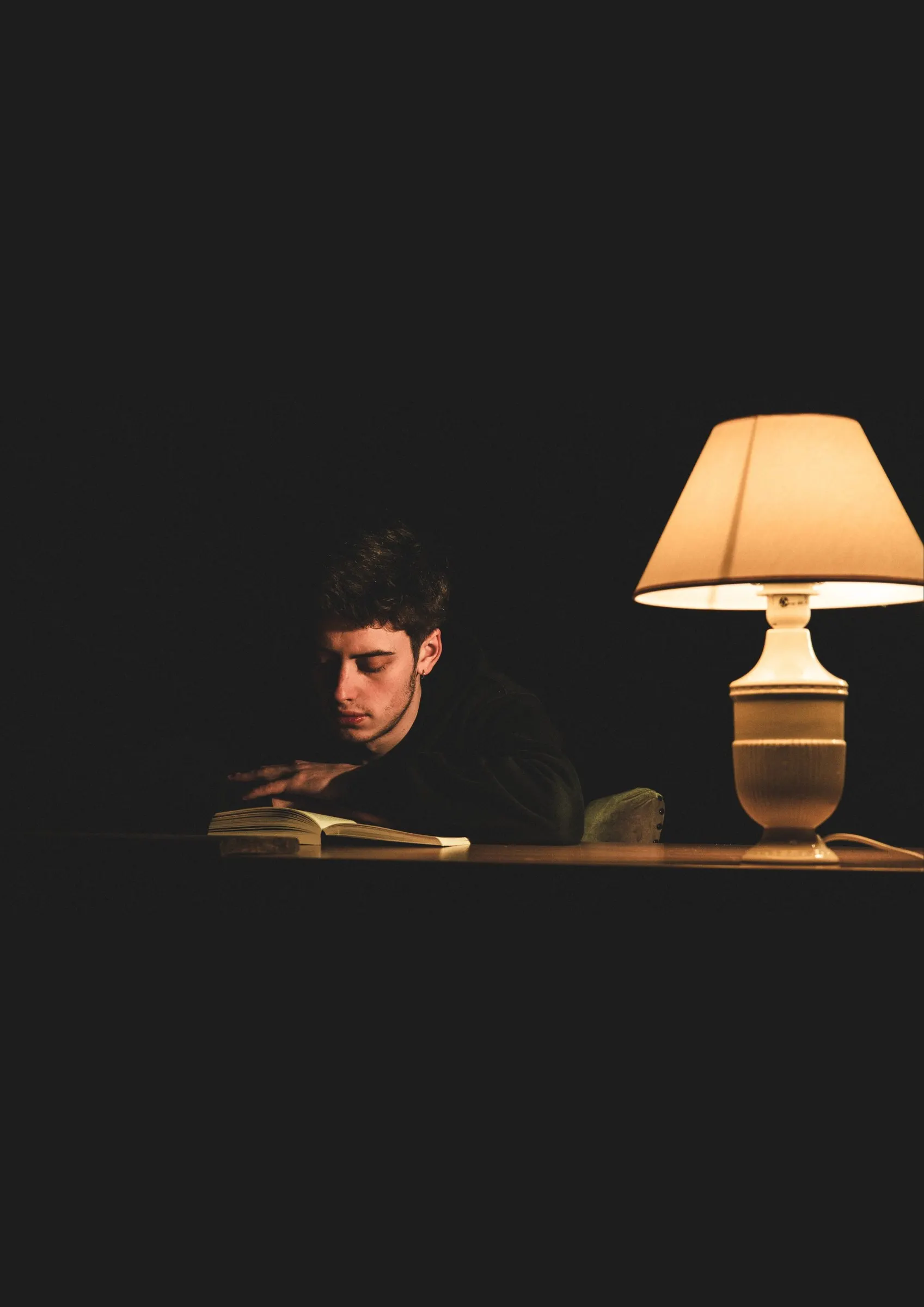 Jonge man die 's avonds aan een tafel leest, alleen verlicht door een schemerlamp
